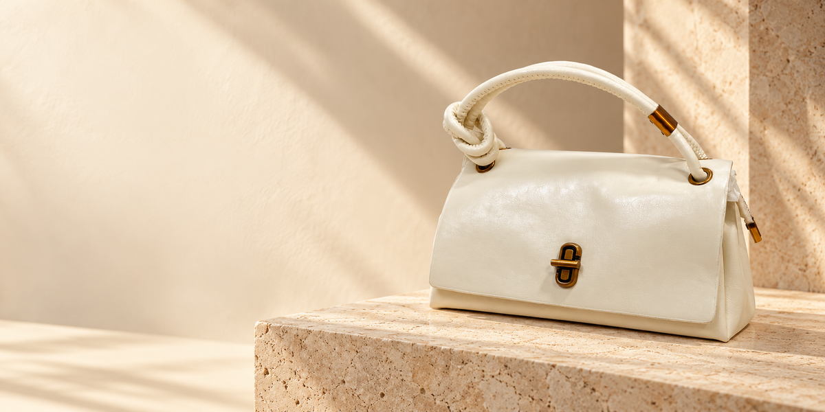 Ivory Surrani pillow bag with knotted top handle and turn-lock clasp on a travertine plinth